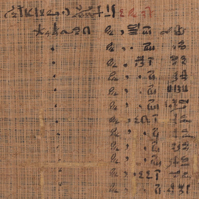 A close view of the tan woven surface of the Ebers Papyrus shows black hieratic writing across the top and in a vertical table at right, with a short passage in red ink near the upper edge.