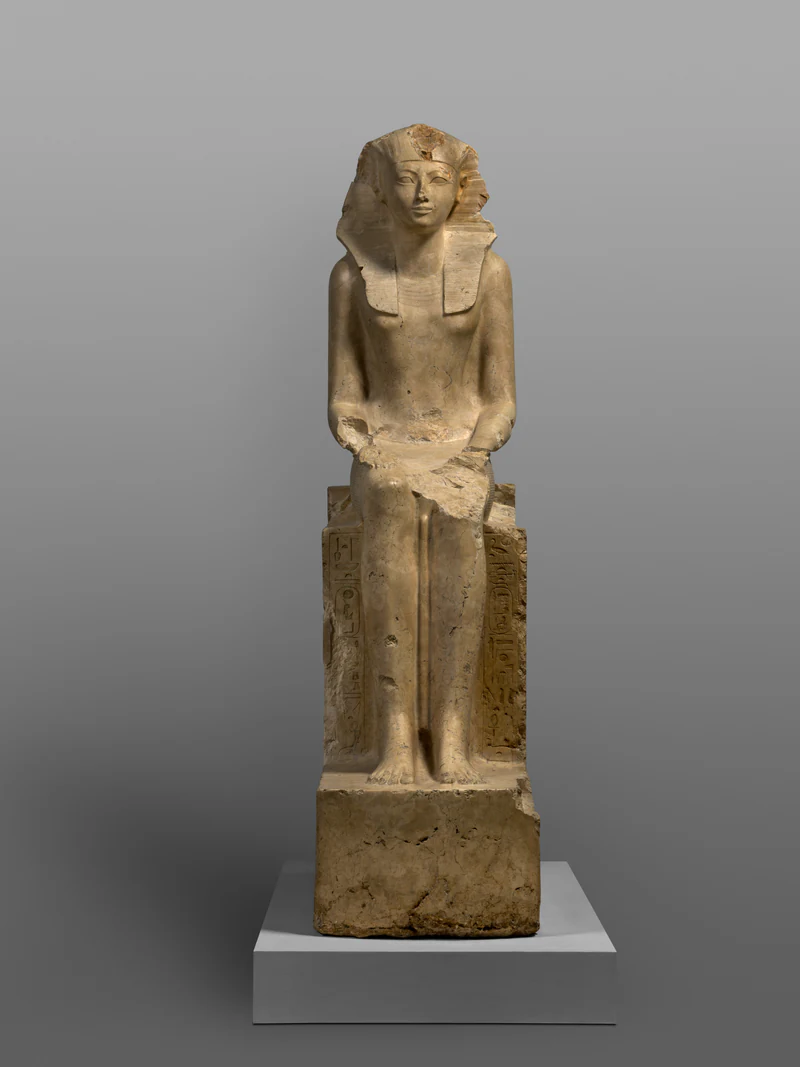 A pale limestone statue of Hatshepsut seated frontally on a throne, wearing a royal nemes headdress. Her hands rest flat on her thighs, her lower left arm and part of the lap are damaged, and vertical hieroglyphic inscriptions flank her legs.